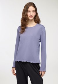 Camicia a maniche lunghe di colore lavanda in tessuto morbido, con scollatura rotonda e orlo scallop. Abbinata a pantaloni neri dal taglio morbido.