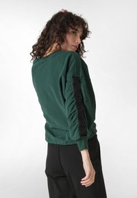 Zelená mikina s volným střihem, žebrovanými manžetami a texturovaným černým logem na rukávu. Měkký materiál s hladkým povrchem.