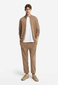 Ung man som bär en beige zip-up kofta, matchande joggerbyxor, vit T-shirt och beige sneakers, står med händerna i fickorna.