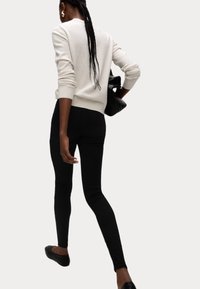Jersey de punto crema con puños acanalados, jeans skinny negros y bailarinas negras. La figura lleva un bolso negro y tiene el cabello largo trenzado.