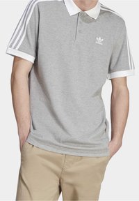 Graues Poloshirt mit weißem Kragen und Bündchen, Dreiknopfverschluss und drei weißen Streifen auf den Schultern. Adidas-Logo auf der linken Brust.