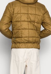 Brun quiltad pufferjacka med huva, med en åtdragen nederkant och elastiska manschetter, kombinerad med vita byxor mot en enfärgad bakgrund.