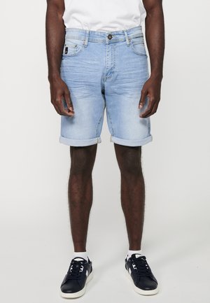 Shorts en denim bleu clair avec des revers, coupe régulière, design à cinq poches et un bouton en métal brillant à la taille. Portés avec des baskets navy.