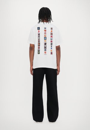 T-shirt blanc à manches courtes avec un design graphique coloré et divers icônes au dos. Associé à un pantalon large noir.