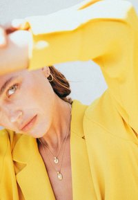 Guldhalsband som är lager på lager med en cirkulär berlock och oval ädelsten, bärs över en skjortblazer i klart gult. Slät textur och elegant design.