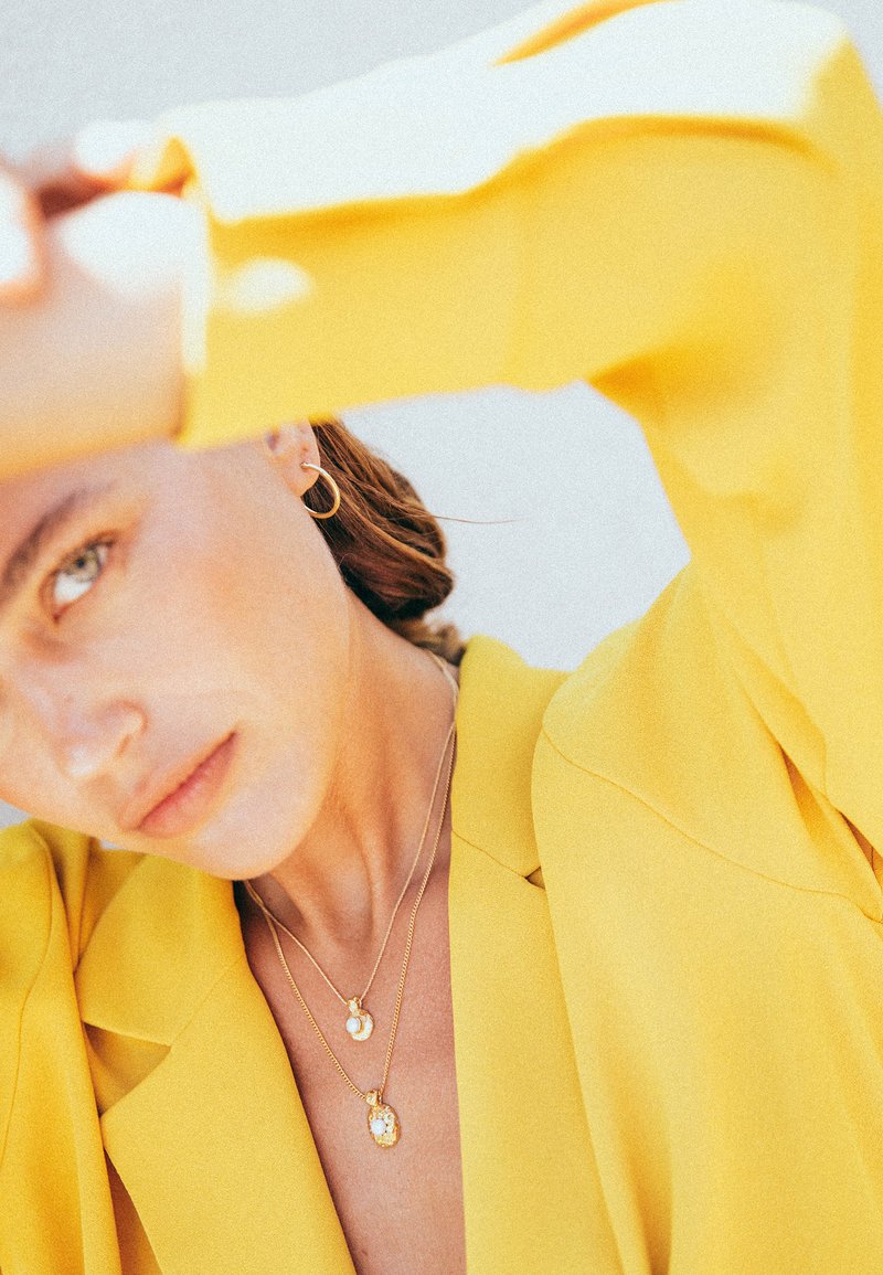 Guldhalsband som är lager på lager med en cirkulär berlock och oval ädelsten, bärs över en skjortblazer i klart gult. Slät textur och elegant design.