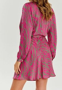 Mujer vistiendo un vestido largo de manga larga con estampado abstracto de rayas de cebra en rosa y verde, vista desde atrás, con el cabello largo y ondulado suelto.