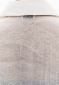 Kremowa koszula z tradycyjny kołnierzykiem, wykonana z lekkiego, pomarszczonego materiału o subtelnej teksturze, z widoczną metką przy dekolcie.