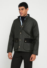 Πράσινο-ελαιί τζάκετ με κερωμένη υφή, διακοσμημένο με καφέ τσέπες από κοτλέ ύφασμα και χάλκινα κουμπιά με πατούρες. Διαθέτει ψηλό γιακά και άνετη εφαρμογή.