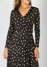 Kobieta ubrana w czarną sukienkę z długim rękawem w białe kropki, z dekoltem w kształcie kopertowym i związanym paskiem w talii, stoi na jednolitym tle.