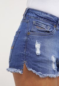 Siniset farkkushortsit kuluneilla yksityiskohdilla. Mukana röpellyt helma, kaksi takataskua ja pienet sivuhaarat alareunassa.