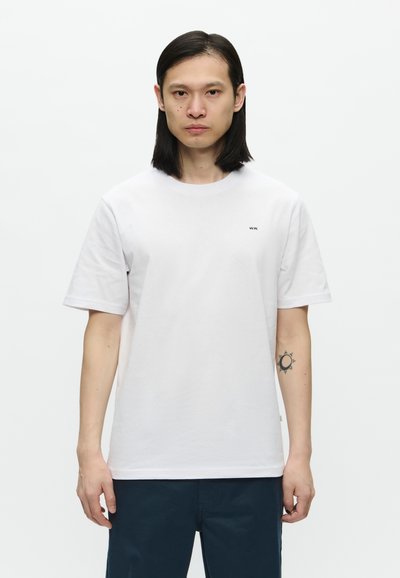 Camiseta blanca de algodón con mangas cortas, cuello redondo y un pequeño logo negro en el pecho. Textura suave y ajuste relajado.