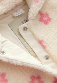 Veste pour bébé de couleur crème douce avec des motifs de fleurs roses, boutons-pression en métal et une étiquette visible portant la mention "NEXT baby Fabriqué en Chine."