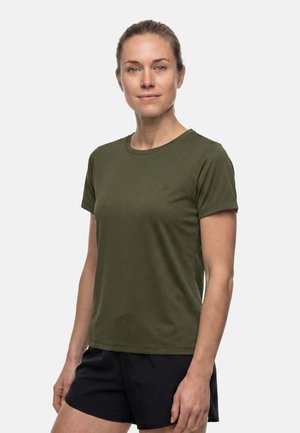 Femme aux cheveux attachés porte un t-shirt manches courtes vert foncé et un short noir, debout devant un fond clair uni.