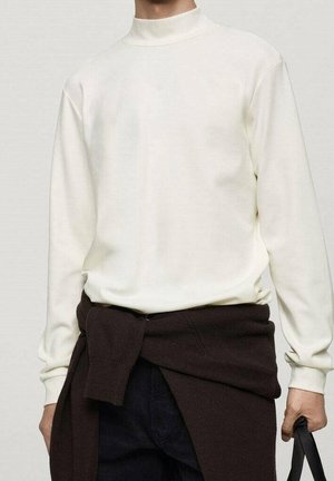Homme portant une chemise blanche à col montant à manches longues avec un pull sombre noué autour de la taille, tenant un sac à bandoulière noir dans la main droite.