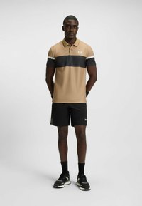 Lysebrun og mørkegrå polo med ternet mønster, korte ærmer og et logo. Kombineret med sorte shorts og sportssko, står lige op.