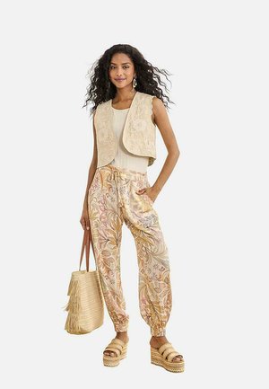 Mujer con cabello largo y rizado lleva un chaleco bordado color crema, pantalones con estampado floral, sandalias de plataforma y sostiene un bolso tejido con borlas.
