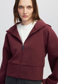 Sudadera corta burdeos con cremallera y capucha, que presenta una textura suave y costuras planas. Combinada con una camiseta blanca de cuello redondo por debajo.