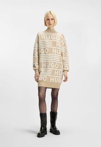 Beige neulemekko, jossa on suuri valkoinen "HUGO" -teksti, korkea kaulus ja pitkät hihat. Yhdistetty mustiin polvipitkiin saappaisiin. Pehmeä rakenne.