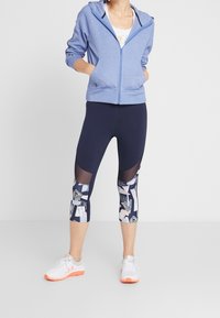 Sudadera de cremallera azul claro hecha de tela suave, combinada con leggings capri azul marino que presentan paneles de malla y patrones abstractos en varios colores. Zapatillas blancas.
