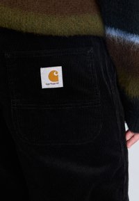 Sorte fløjlsbukser med en baglomme, der har et firkantet hvidt mærke med et stiliseret orange logo og navnet "carhartt."