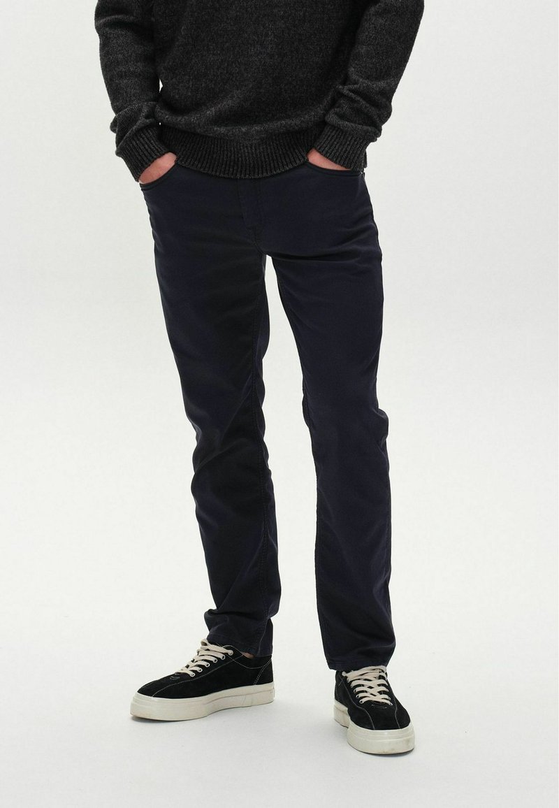 Dunkelblaue Chinos mit schlanker Passform, kombiniert mit schwarzen Wildledersneakern mit weißen Gummisohlen und cremefarbenen Schnürsenkeln. Strukturiertes Strickpullover.