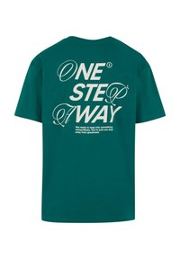 Camiseta de algodón verde con gran texto blanco en la parte trasera: "UN PASO ALEJADO." Textura suave, mangas cortas y un ajuste relajado.