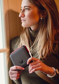 Black leather wallet with a red zipper detail. Square shape, embossed logo on the front, held in a woman's hands.