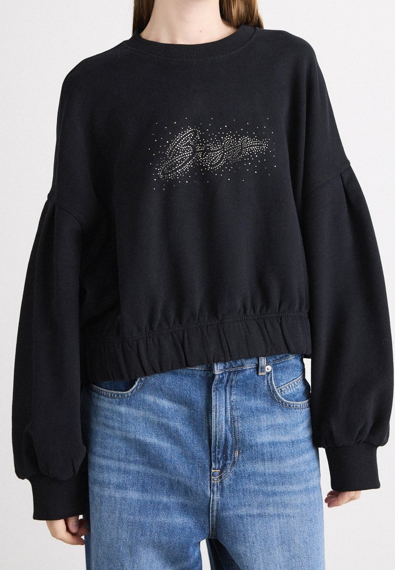 Sweat-shirt court noir à manches longues bouffantes avec détail de texte en strass, associé à un jean bleu taille haute.