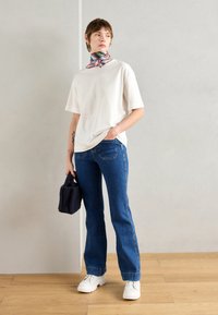 Vit bomullströja, blå utsvängda jeans, vita plattformssneakers och en färgglad silkscarf. Liten mörk handväska syns. Minimalistisk bakgrund.