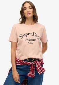 Camiseta de algodón rosa con texto impreso en negro "SuperDry J'adore Paris", combinada con jeans azules y una camisa de cuadros rojos atada a la cintura.