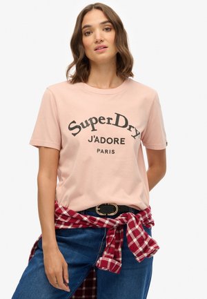 T-shirt en coton rose avec le texte noir imprimé "SuperDry J'adore Paris", associé à un jean bleu et une chemise à carreaux rouges nouée autour de la taille.
