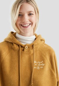 Uśmiechnięta blondynka w musztardowej szmacie z haftem "Bądź miły i szczęśliwy, założone w 1994" oraz białym golfem pod spodem.
