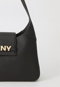 Melnas teksturētas ādas plecu soma ar platu siksnu un zeltainu DKNY logotipu uz priekšējās atloka pret vienkāršu gaišu fonu.