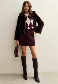 Bruine nepbont jas, wit en bordeaux argyle trui, bordeaux corduroy rok, kniehoge bruine laarzen en een kleine bordeaux handtas.