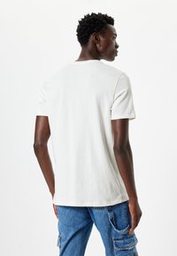 Witte katoenen t-shirt met een geribbelde ronde hals en korte mouwen, gezien vanaf de achterkant. Het model combineert het met blauwe cargobroek.