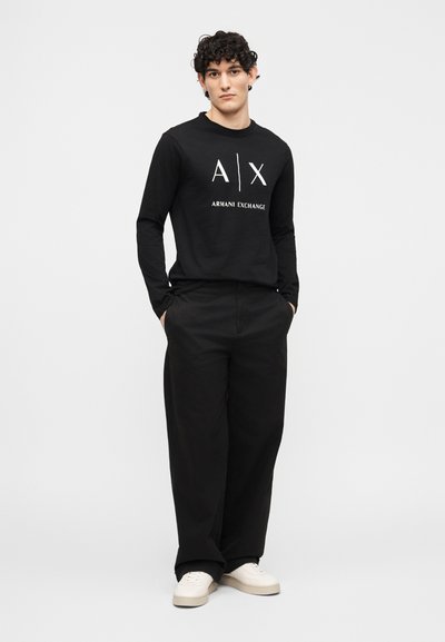 Млад мъж с къдрави коси, облечен с черна дълга риза Armani Exchange, свободни черни панталони и бели маратонки, стои пред едноцветен фон.