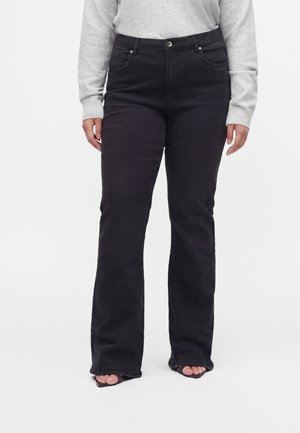 Kvinde iført sorte jeans med høj talje og en smule vidde, kombineret med en lysegrå langærmet top og åbne sandaler.