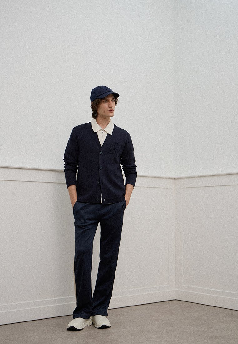 Marinerblauer Cardigan mit weißem Kragen, ausgestattet mit Knopfverschluss und gesticktem Detail, kombiniert mit passenden marinerblauen Hosen und einer Mütze. Weiße Sneakers.