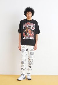 Camiseta negra de gran tamaño con una impresión gráfica colorida de Michael Jordan, combinada con pantalones de chándal blancos con un patrón y zapatillas grises.