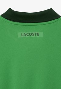 Grön polotröja med en mörkgrön ribbad krage och en rektangulär etikett med "LACOSTE" tryckt i fet svart text. Tillverkad av slät tyg.