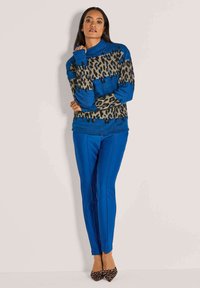 Frau, die an einer Wand steht und einen blauen Leopardenmuster-Pullover, leuchtend blaue Hosen und spitze Leopardenmuster-High Heels trägt.