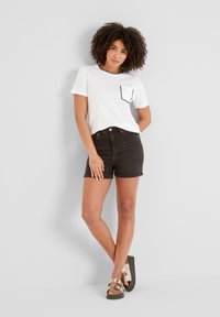 Woman with curly hair stands against white wall wearing white short-sleeve top with black-trimmed pocket, black shorts, and metallic sandals.