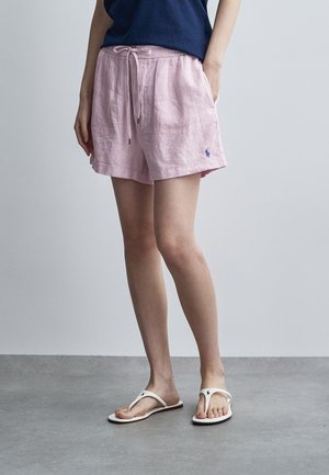 Persoon draagt lichtroze shorts met trekkoord, een marineblauw topje en witte slippers, staand op een grijze vloer tegen een effen grijze muur.