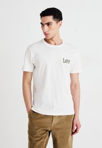 Hvit bomull T-skjorte med en liten grønn "Lee" logo på brystet, sammen med beige kordfløyelsbukser. En enkel, uformell design.