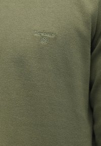 Κοντινής απόστασης εικόνα από πράσινο πλεκτό ύφασμα με το λογότυπο "Barbour" κεντημένο με αντίστοιχο νήμα στην περιοχή του στήθους.