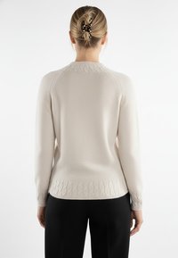 Kremowy sweter z prążkowanym kołnierzem, teksturowanym dołem i akcentami w formie splotu warkocza. Długie rękawy i dopasowany fason, w zestawieniu z czarnymi spodniami.