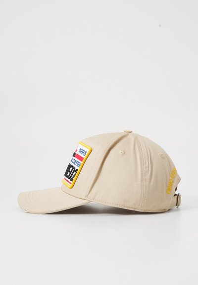 Casquette de baseball en tissu beige avec une visière courbée. Présente un écusson brodé multicolore et du texte sur le côté en jaune. Sangle ajustable à l'arrière.