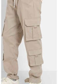 Beige cargobroek gemaakt van zachte stof, met meerdere grote zakken, verstelbare enkels en een gestikt merklogo op de zijzak.
