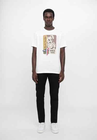 Tricou alb din bumbac, cu un grafic al unui câine din desene animate, text negru și accente geometrice. Asortat cu pantaloni negri și adidași albi.
